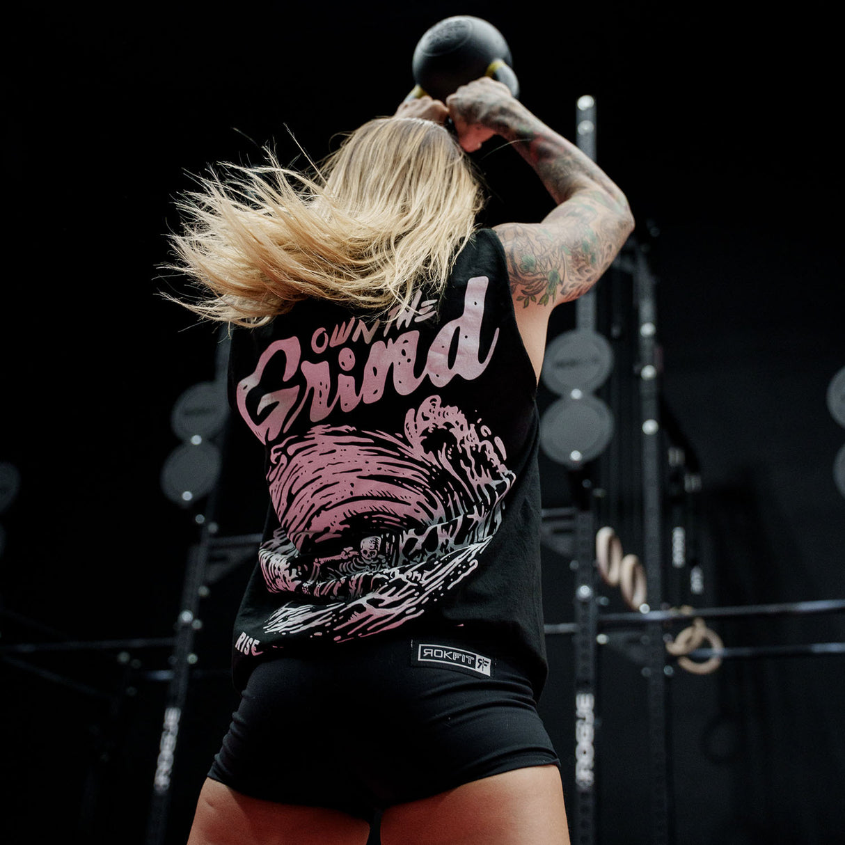 Female model doing a kettlebell swing in a gym wearing sleeveless Street Tee with 'Own The Grind' graphic
