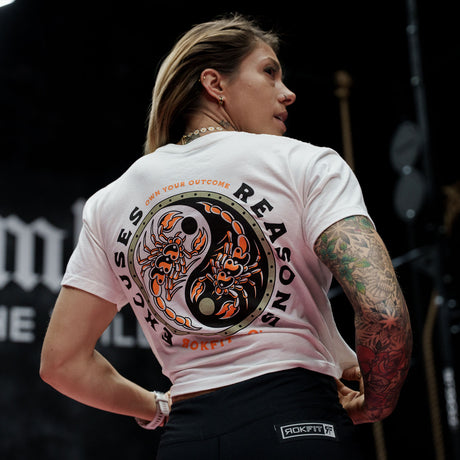 Female model in a gym wearing white crop top with 'Excuses or Reasons' graphic